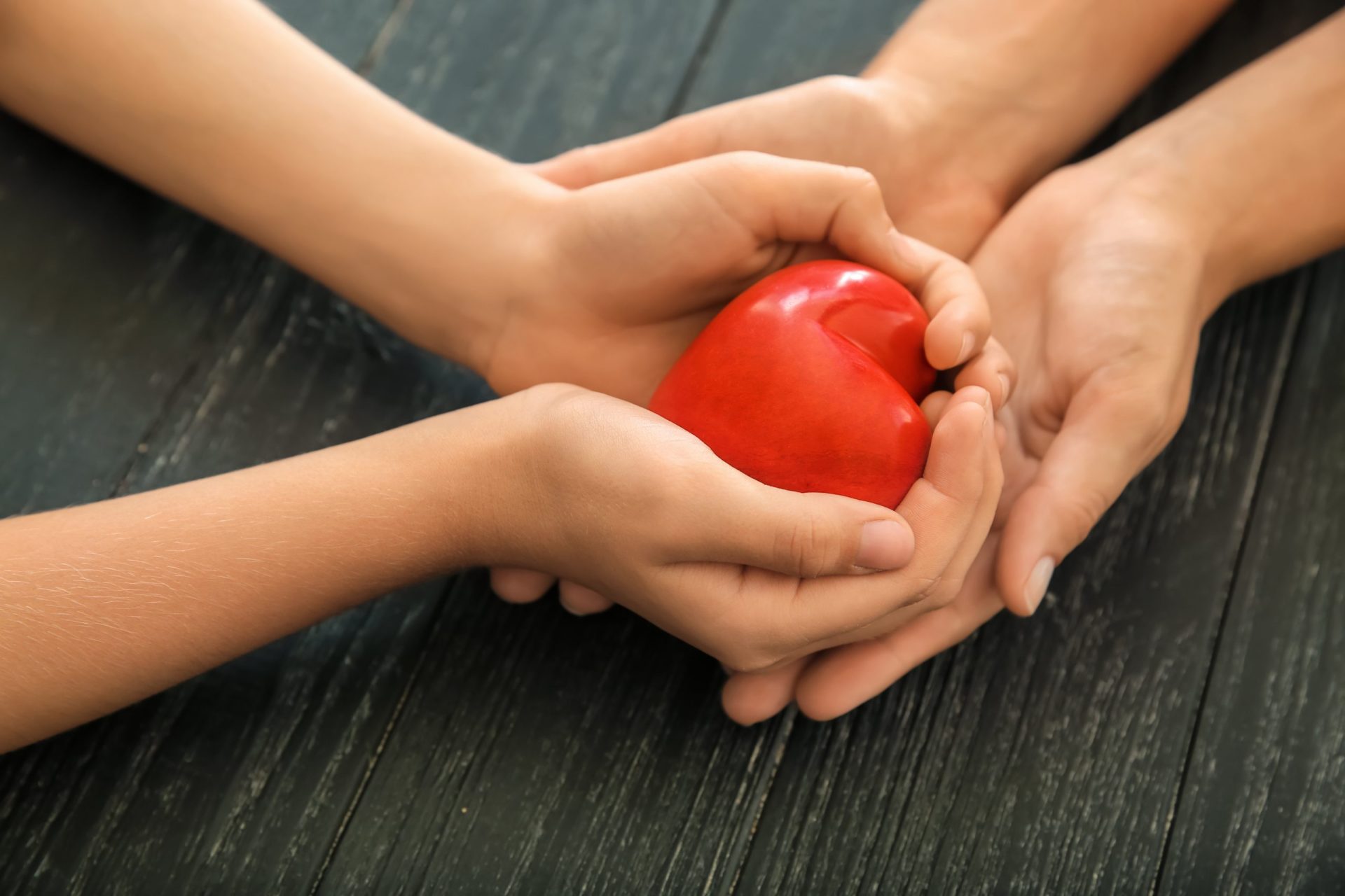 Hands Of Mother And Child Holding Red Heart On Wooden Background